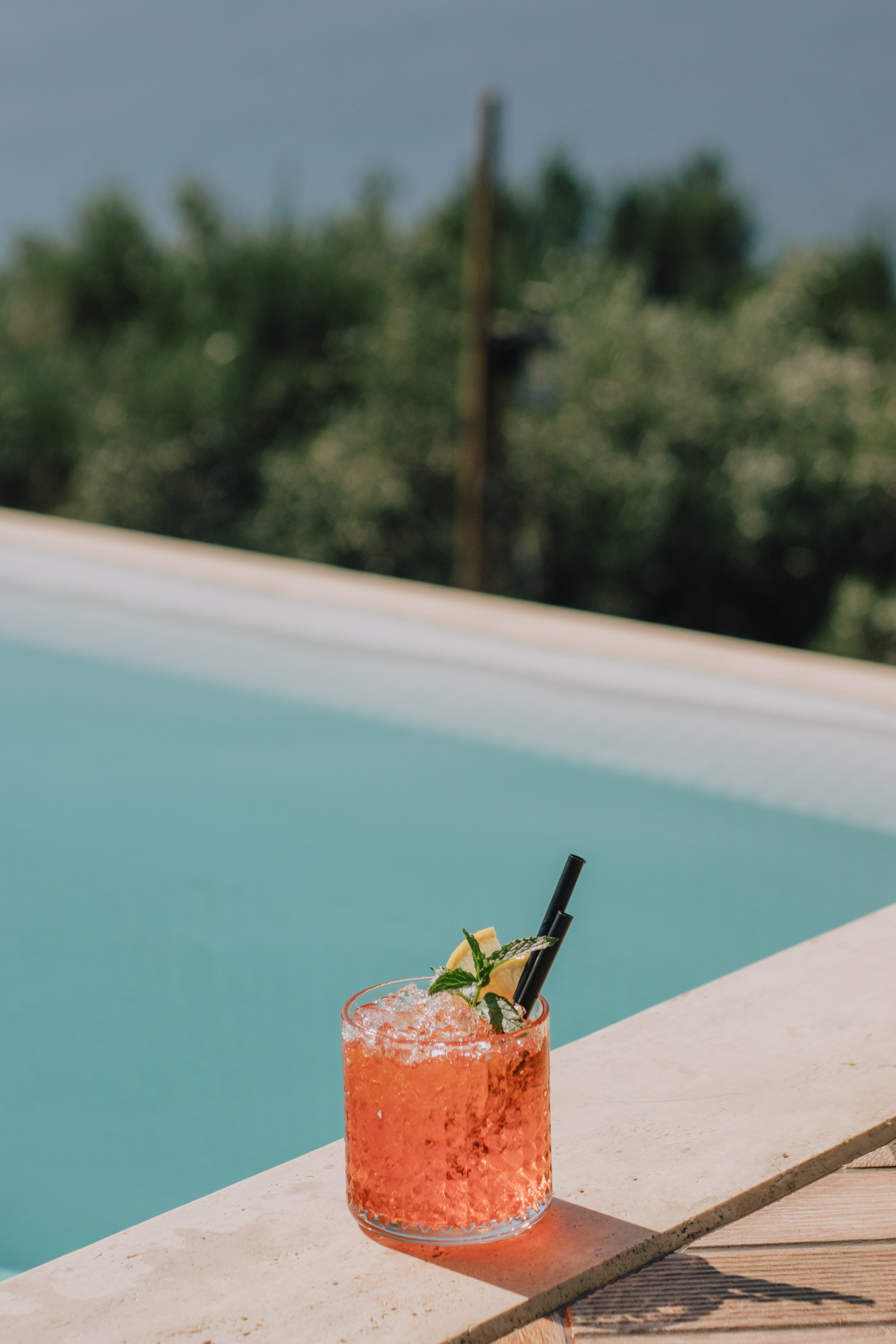 A pink iced drink with mint and citrus garnish resting on the edge of a pool on a sunny day.