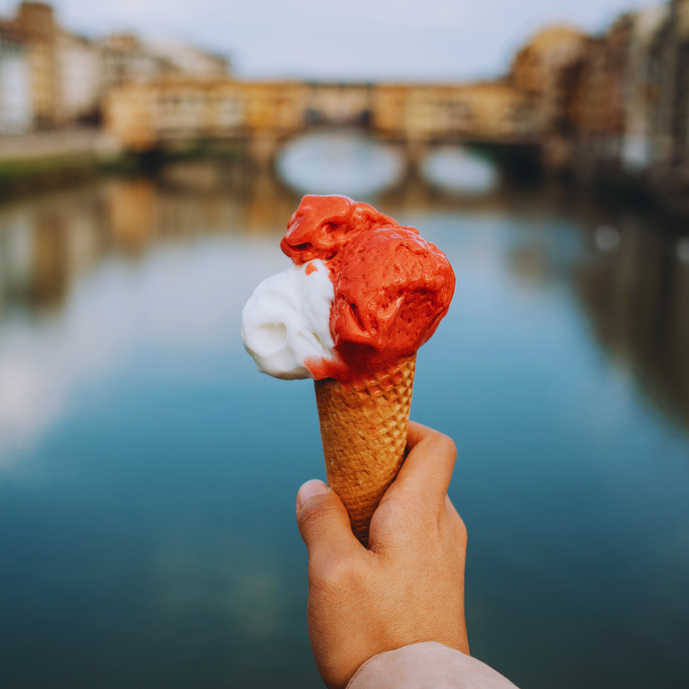 Hand holding a waffle cone with red and white ice cream, held up against a calm river and blurred bridge in the background.