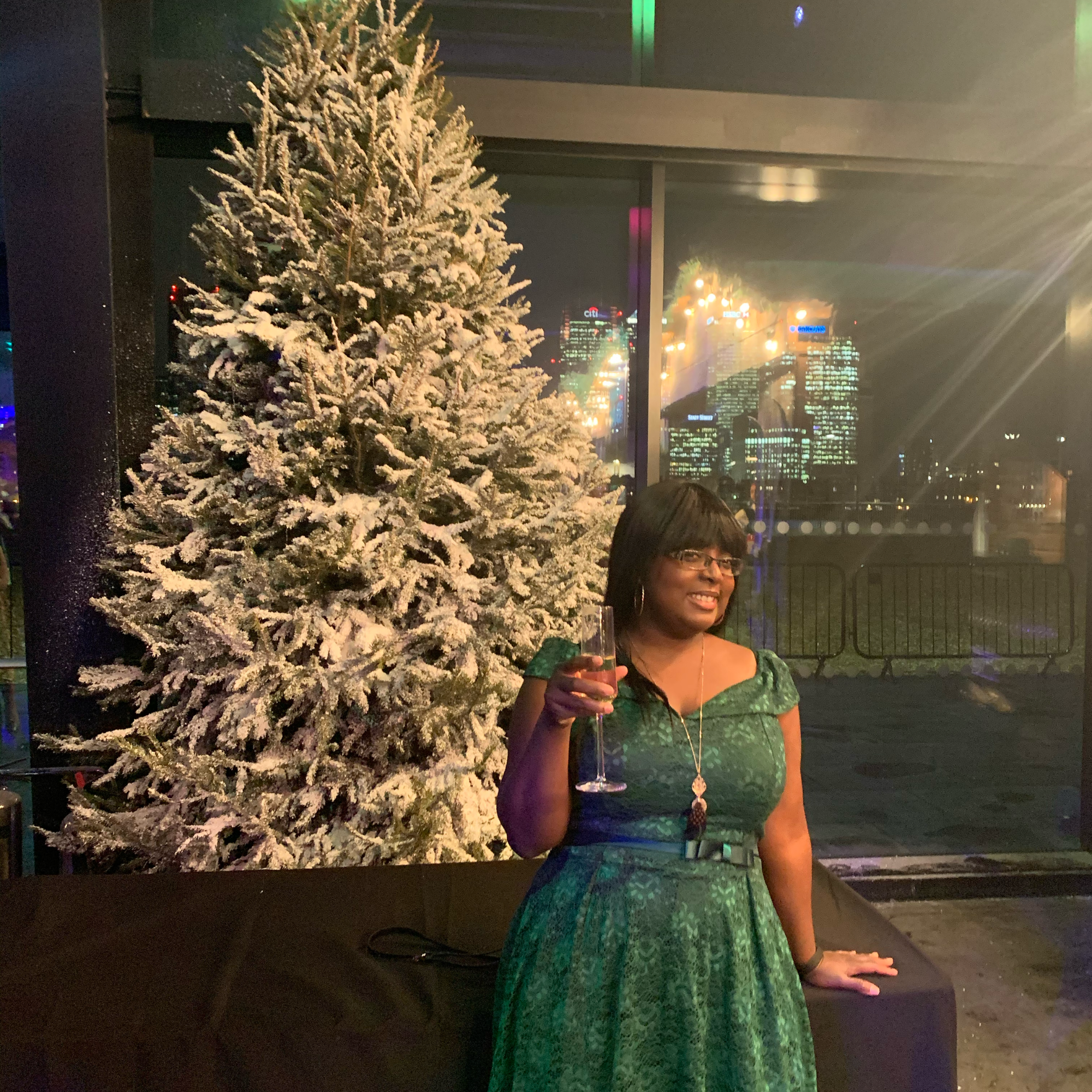 Nefe holding a drink and smiling beside a decorated Christmas tree at night, with city lights in the background.
