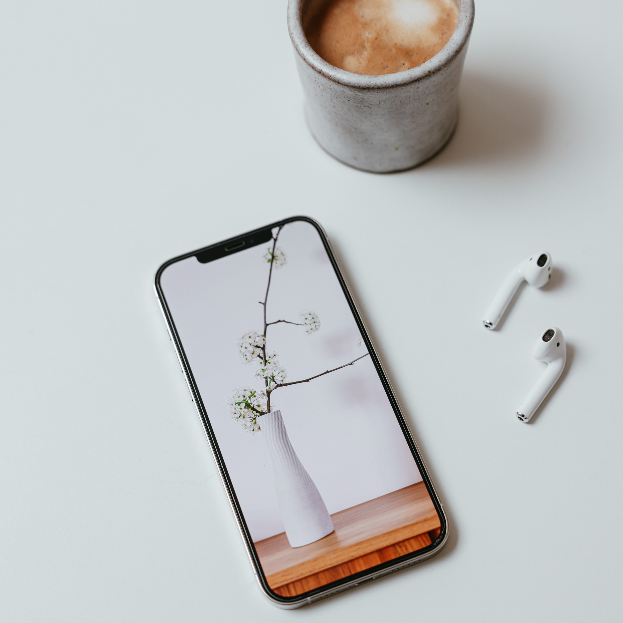 A smartphone displaying a minimalist floral image beside a cup of coffee and wireless earbuds on a white surface.