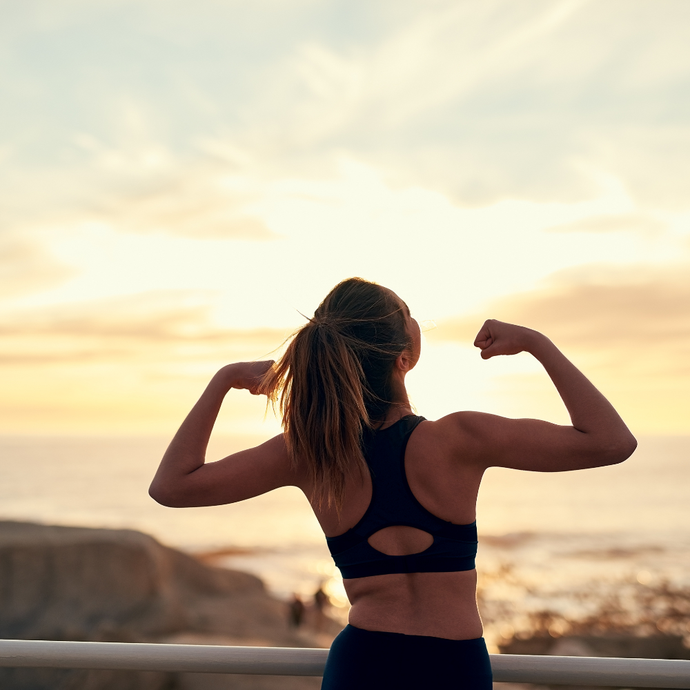 10 Build Success with Effective Daily Habits A woman in athletic wear flexing her arms triumphantly while facing a sunrise by the seaside.