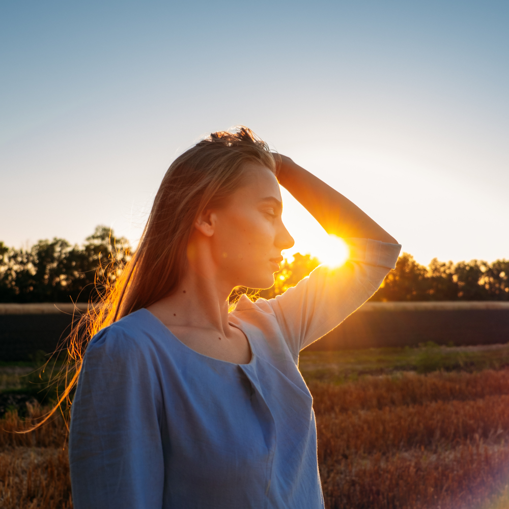 A woman standing in a field at sunset, with the sun shining through her hair as she holds her hand to her head.