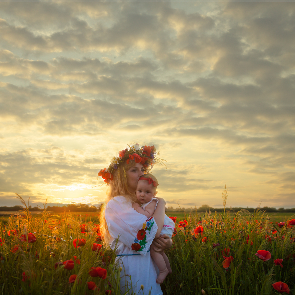 A mother wearing a flower crown holds her baby in a field of red poppies at sunset, symbolising the nurturing and growth that come from setting inspiring goals.