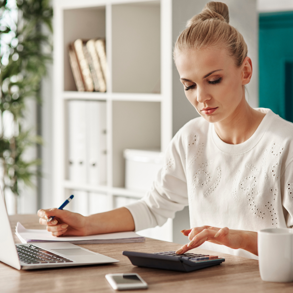 11 How To Master Smart Budgeting A focused woman calculating finances at her home office, using a calculator and laptop.