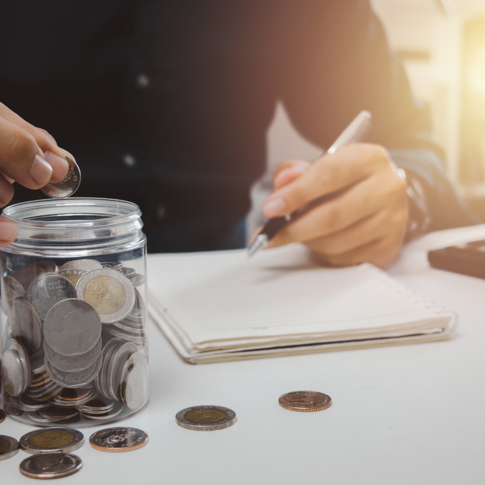 Person dropping coins into a jar while writing in a notebook, symbolising financial planning and investment.