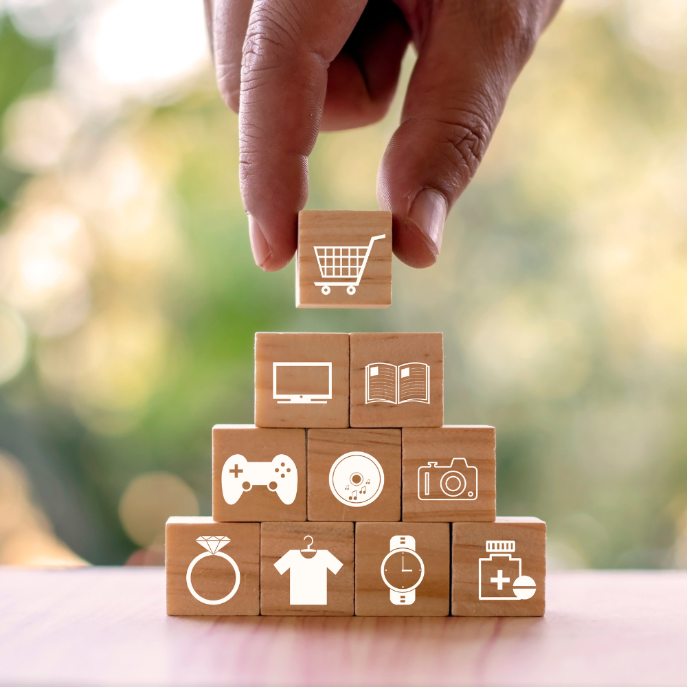 Hand placing a wooden block with a shopping cart icon on top of a pyramid of blocks featuring various icons representing different consumer goods and services.