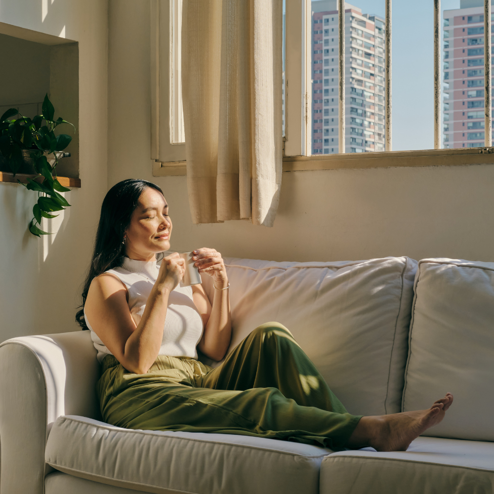 18. Mindfulness Techniques for Professionals Woman practising mindfulness by relaxing on a sofa with a cup of tea in a sunlit room.