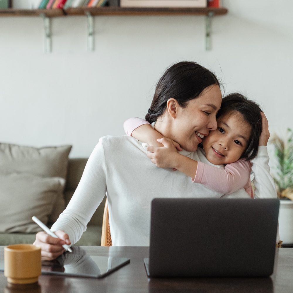 19. Top Tips for Balancing Work and Relationships A smiling woman working on a laptop with a stylus while her young daughter hugs her from behind, symbolising the balance between work and family life.