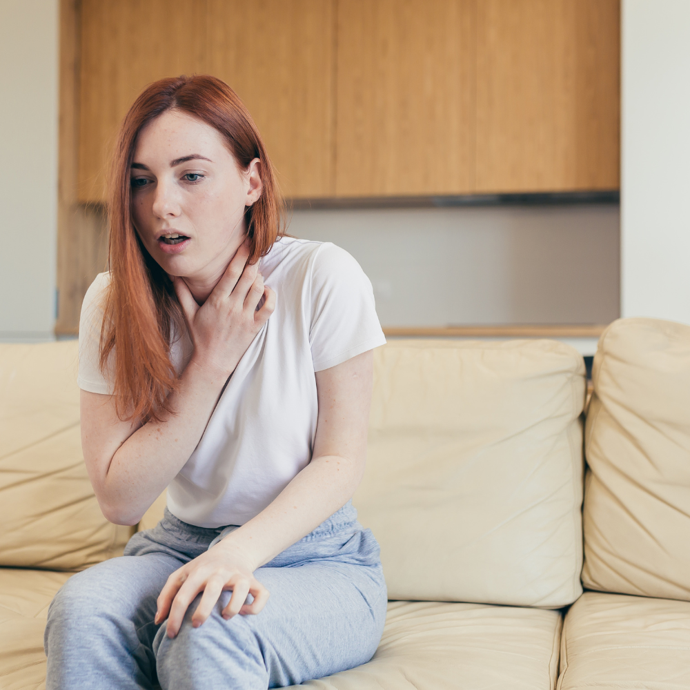 A woman experiencing a panic attack, holding her neck and looking distressed while sitting on a beige sofa in a modern living room.