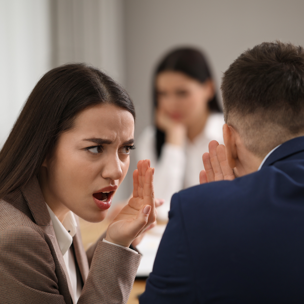 21. Overcoming Workplace Bullying A Guide to Reclaiming Your Confidence Two colleagues whispering and gossiping at work while a concerned woman looks on.