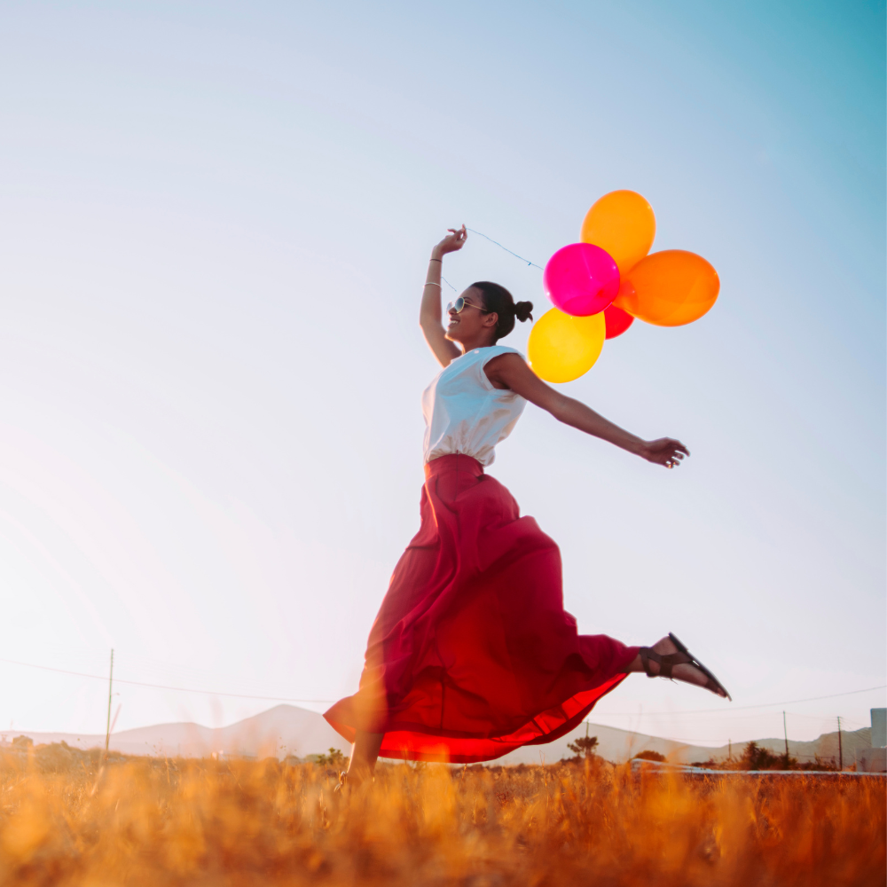 Woman running joyfully through a field while holding colourful balloons under a clear sky.