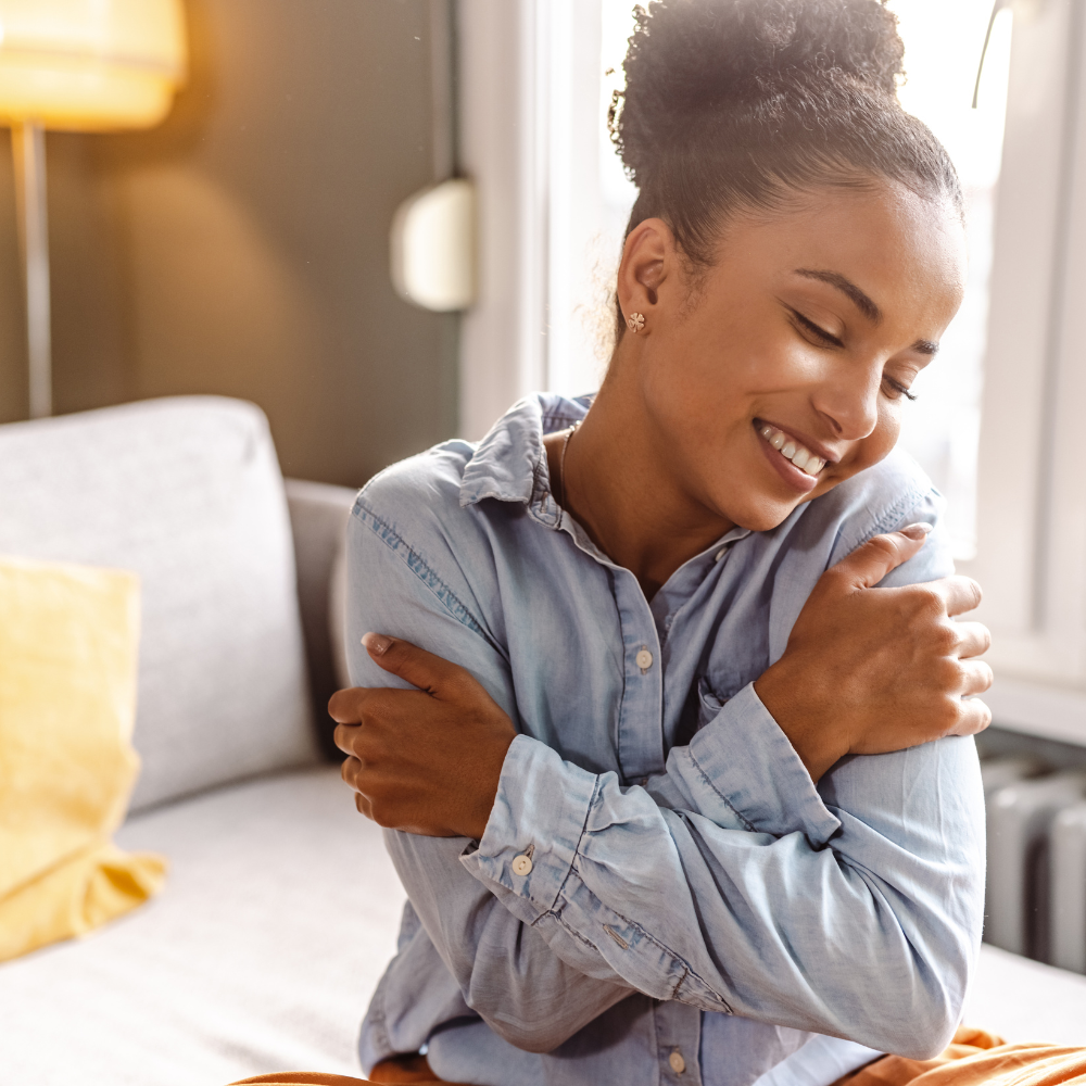 A joyful woman embracing herself with a warm smile, symbolising self-love, inner peace, and self-acceptance in a cosy, light-filled room.