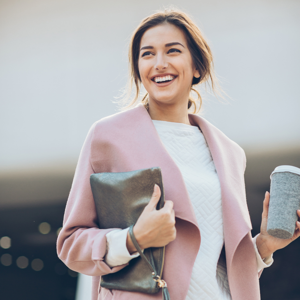 24. The Confidence Catalyst Unlocking Your Potential in the Final Quarter Confident woman smiling while holding a coffee cup and clutch bag, wearing a pink coat, symbolising self-assurance and readiness to take on new challenges.
