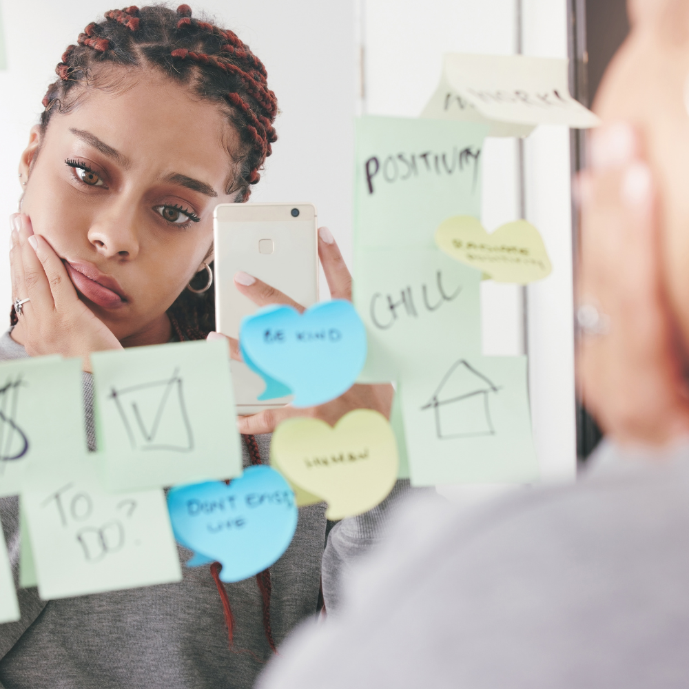Woman reflecting in the mirror, surrounded by post-it notes with positive affirmations.