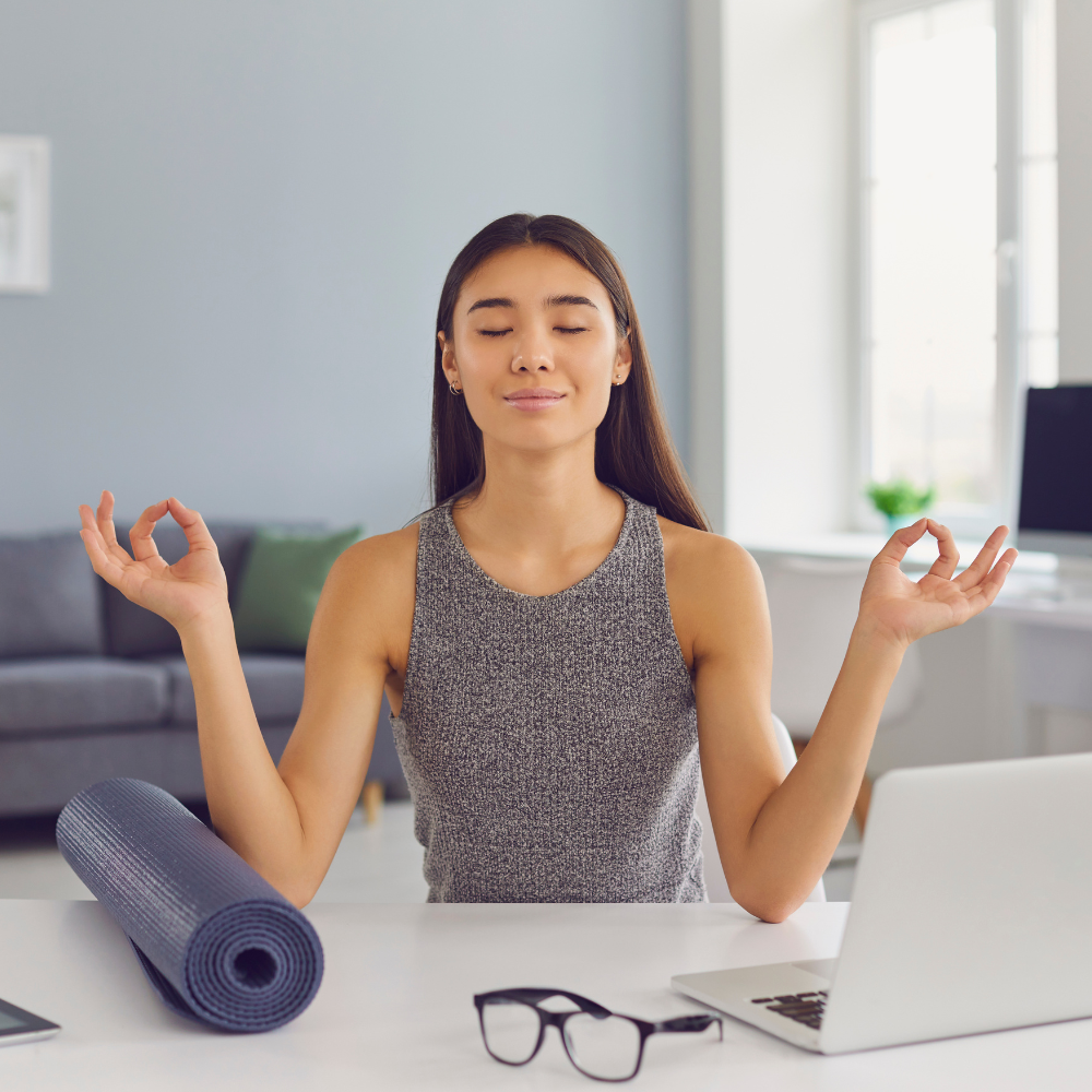 Woman practicing mindfulness at her desk with closed eyes and a yoga mat beside her, embodying calm and balance in a modern workspace.