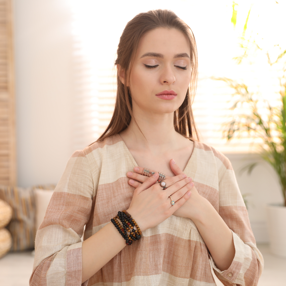 Woman practising self-care by meditating with her hands over her heart, eyes closed in a peaceful setting.