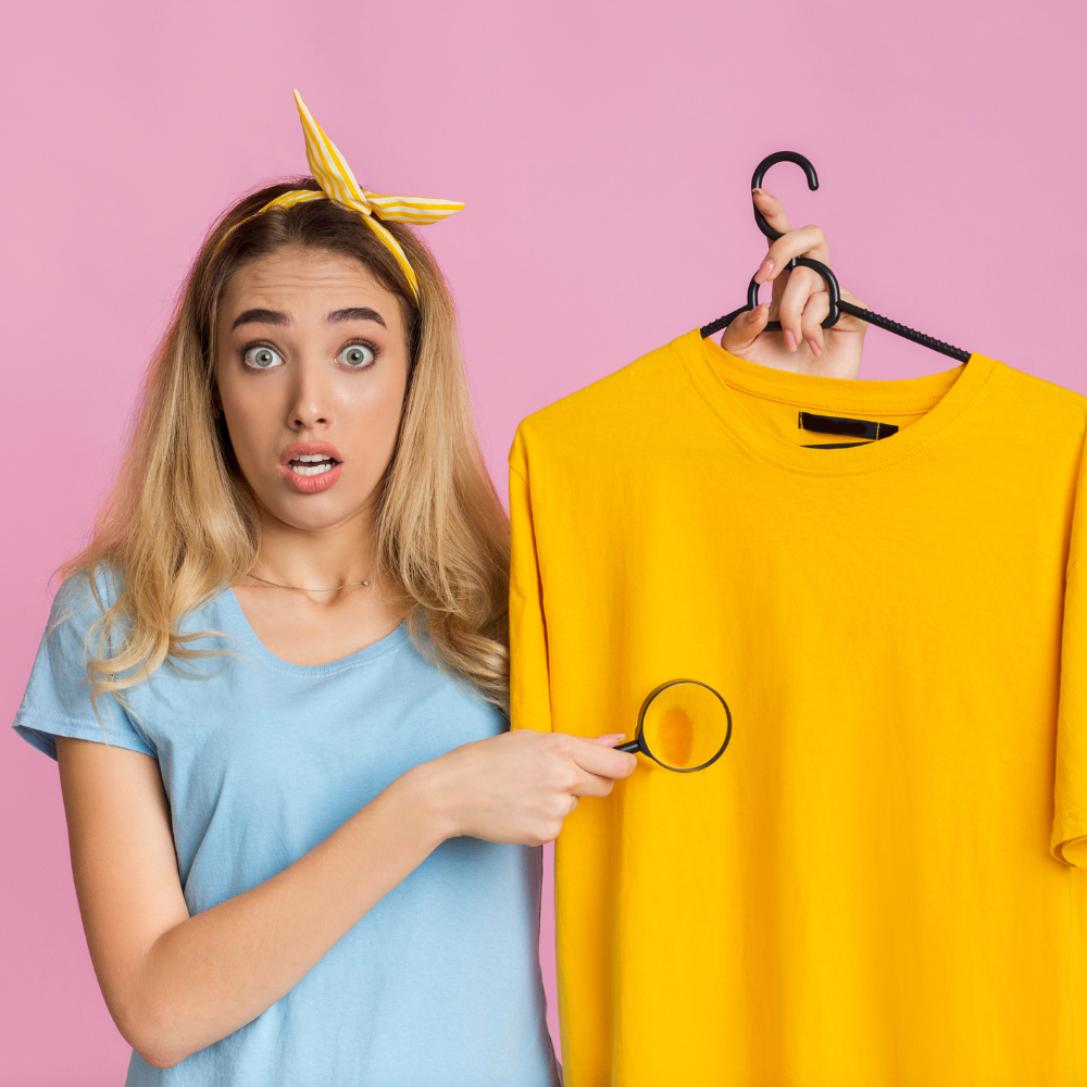 Woman with surprised expression holding a magnifying glass up to a yellow t-shirt, illustrating the concept of perfectionism.