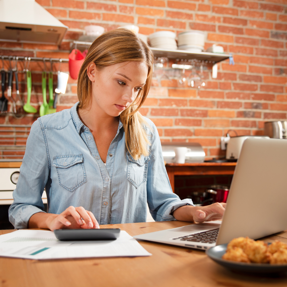 35. Budgeting for Success Women and Money Management A young woman in a casual denim shirt working on her budget at the kitchen table with a calculator and laptop, symbolising financial planning and empowerment.