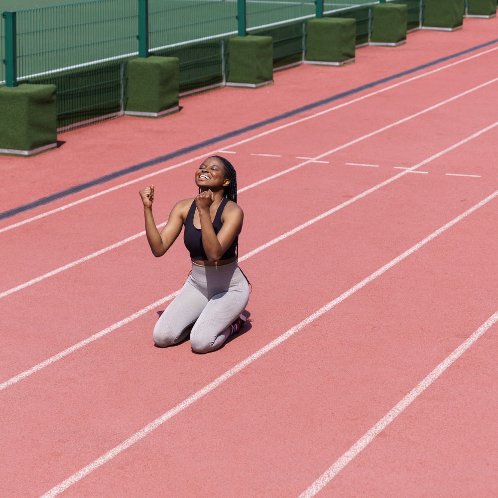 A smiling woman kneels on a running track, raising her fists in a joyful gesture of celebration.