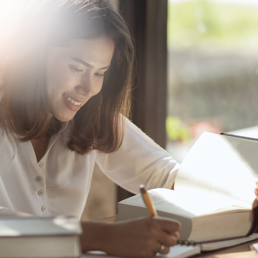 A smiling woman in a bright, sunlit space, writing notes in a notebook while surrounded by open books.