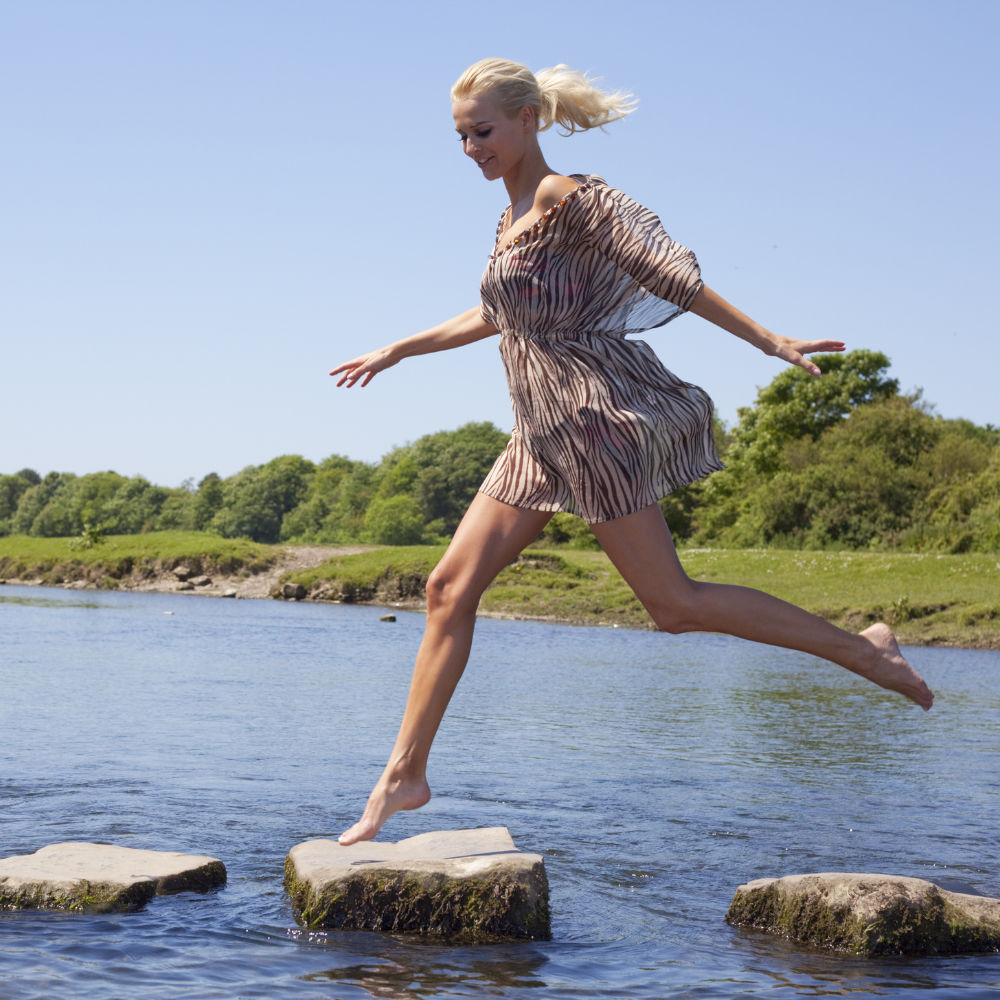 A confident, smiling woman in a light, patterned dress stepping across stones over a calm stream on a sunny day, surrounded by lush green trees.