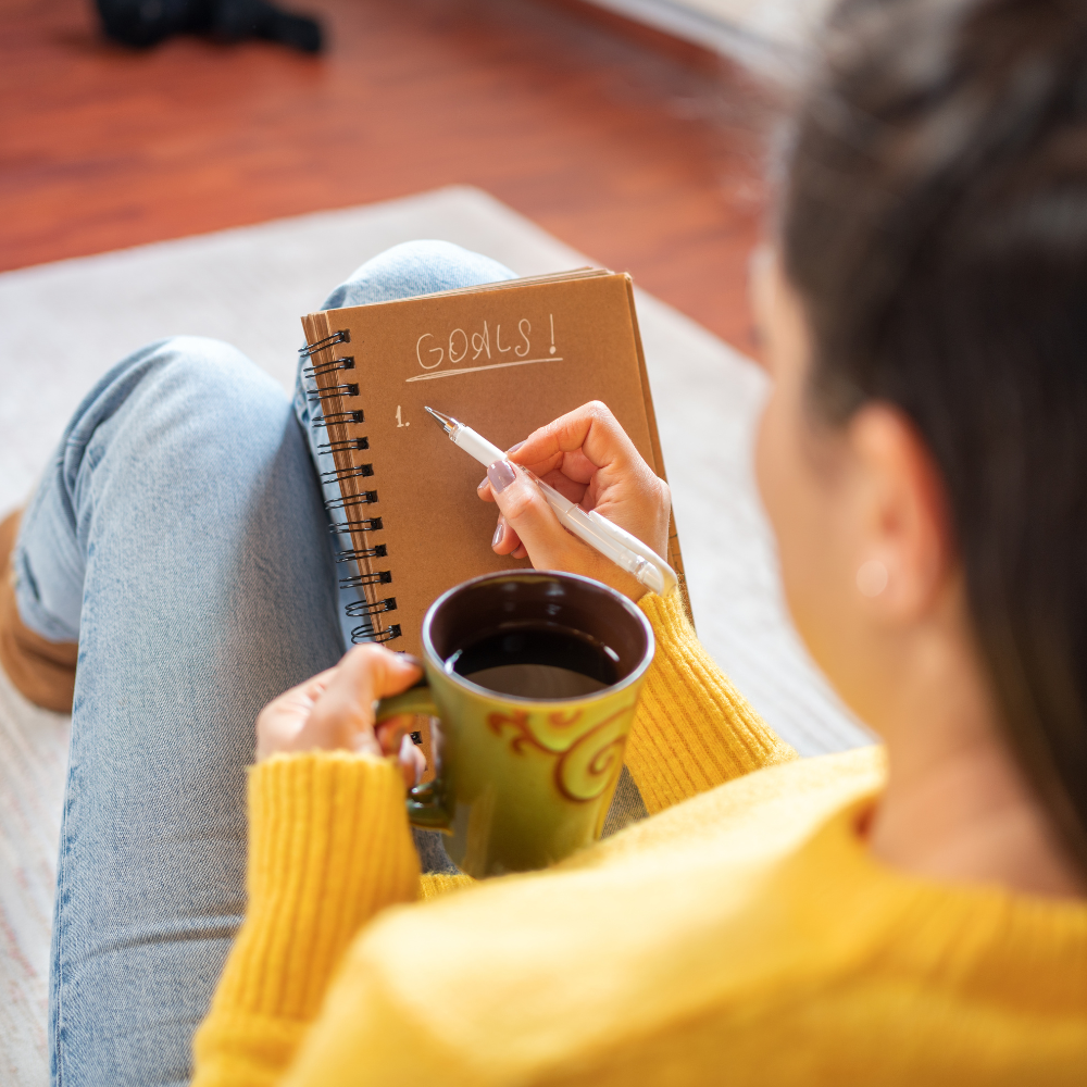 41. From Vision to Action Turning Your New Year’s Goals into Reality A woman in a bright yellow jumper holds a coffee mug while writing “Goals!” in a spiral notebook.