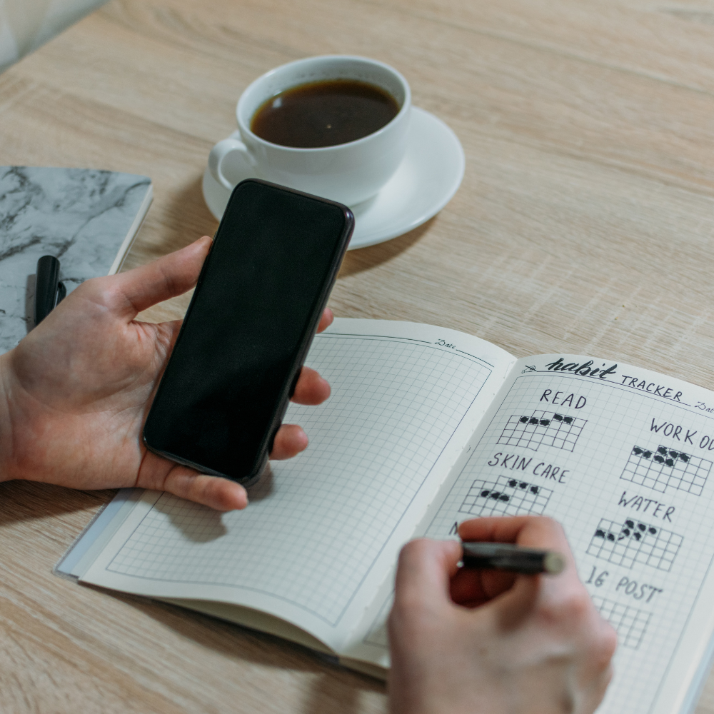 A person holding a smartphone in one hand and marking entries in a habit tracker notebook, with a cup of coffee on the table.