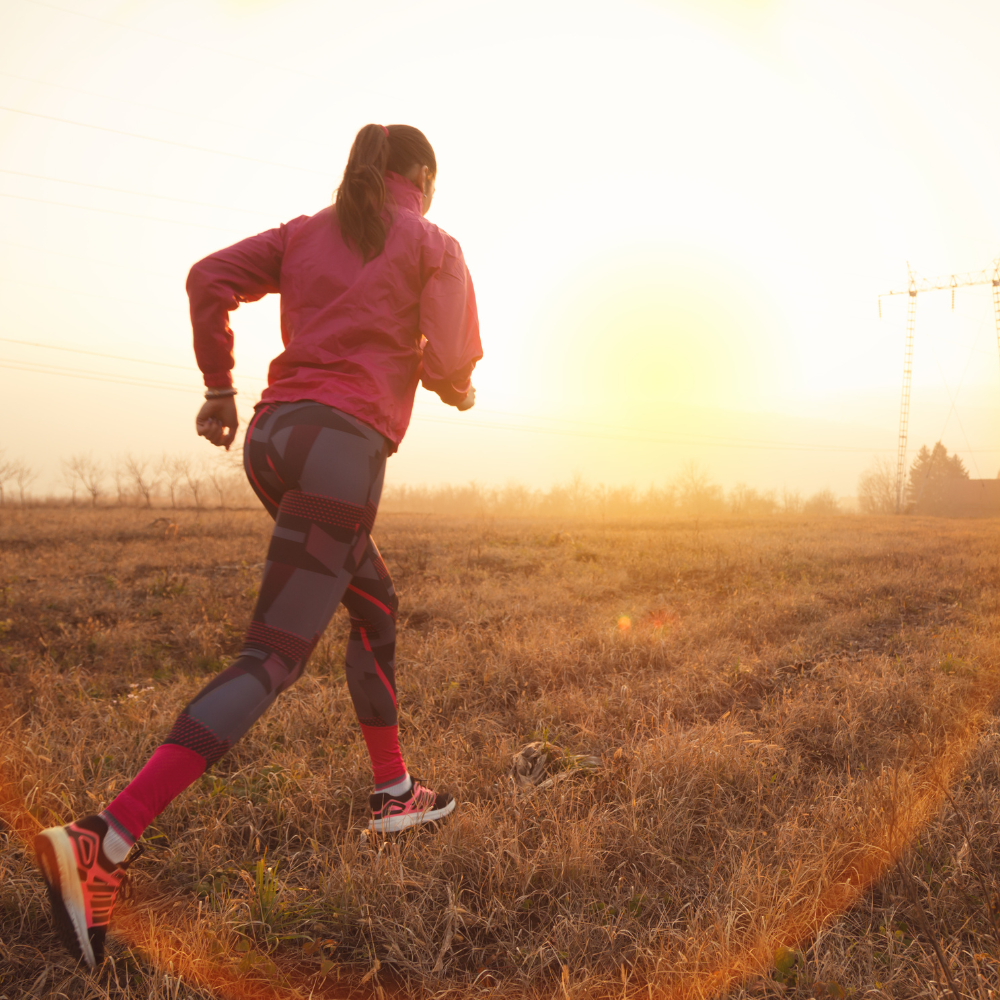 A woman running outdoors in a field at sunrise, wearing a pink jacket and patterned leggings, symbolising motivation and perseverance.