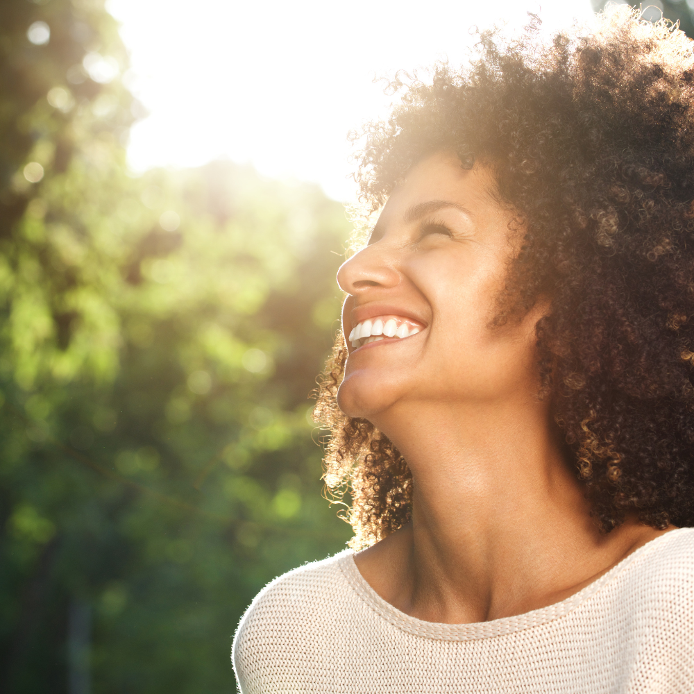 A smiling woman with curly hair looks upwards in bright sunlight, radiating joy against a lush green background.