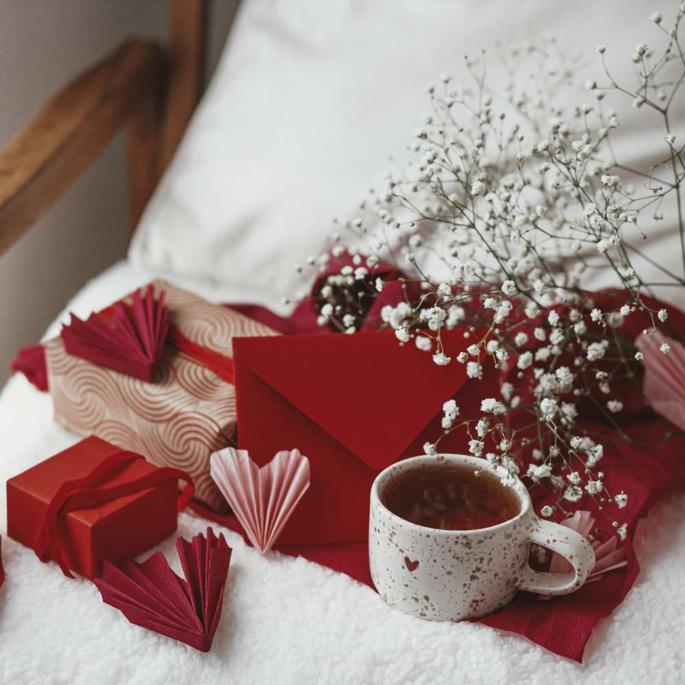 A cosy Valentine’s Day setup with a heart-decorated mug of tea, red envelopes, gift boxes, paper hearts, and white baby’s breath flowers on a soft bed.
