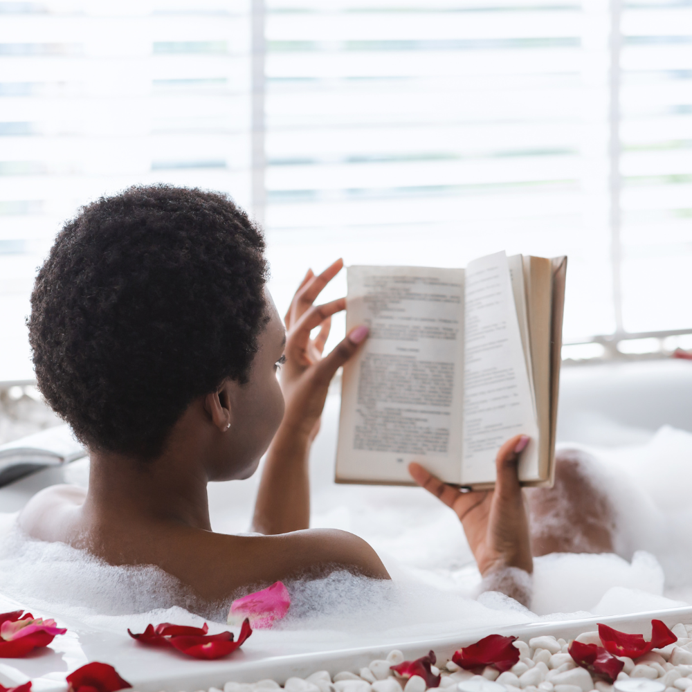 48. Confidence in Self-Care Boundaries & Kindness That Transform Lives A woman with short, natural hair relaxes in a bubble bath filled with rose petals, reading a book by the light of a nearby window.