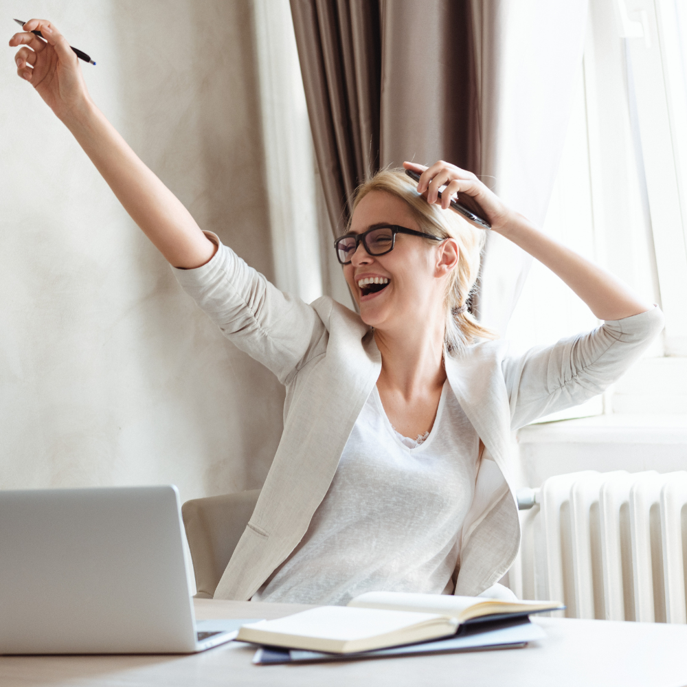 49. Reclaim Your Worth Celebrate Small Wins and Crush March Goals A smiling woman wearing glasses raises one arm in celebration while sitting at a desk with a laptop and an open notebook.