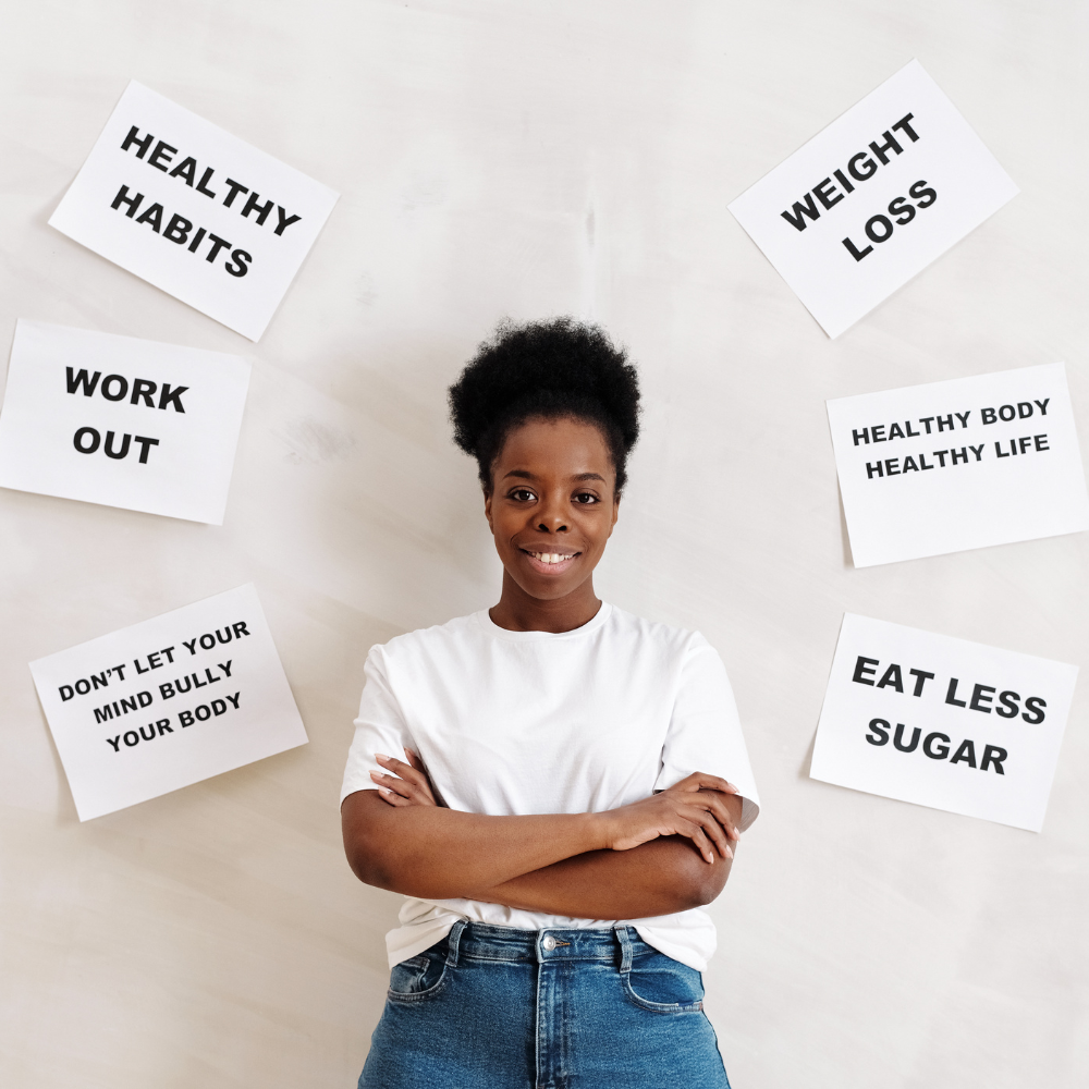 51. Building Empowering Habits for Confidence A smiling Black woman wearing a white T-shirt stands confidently with folded arms, surrounded by motivational posters about healthy habits and body positivity.