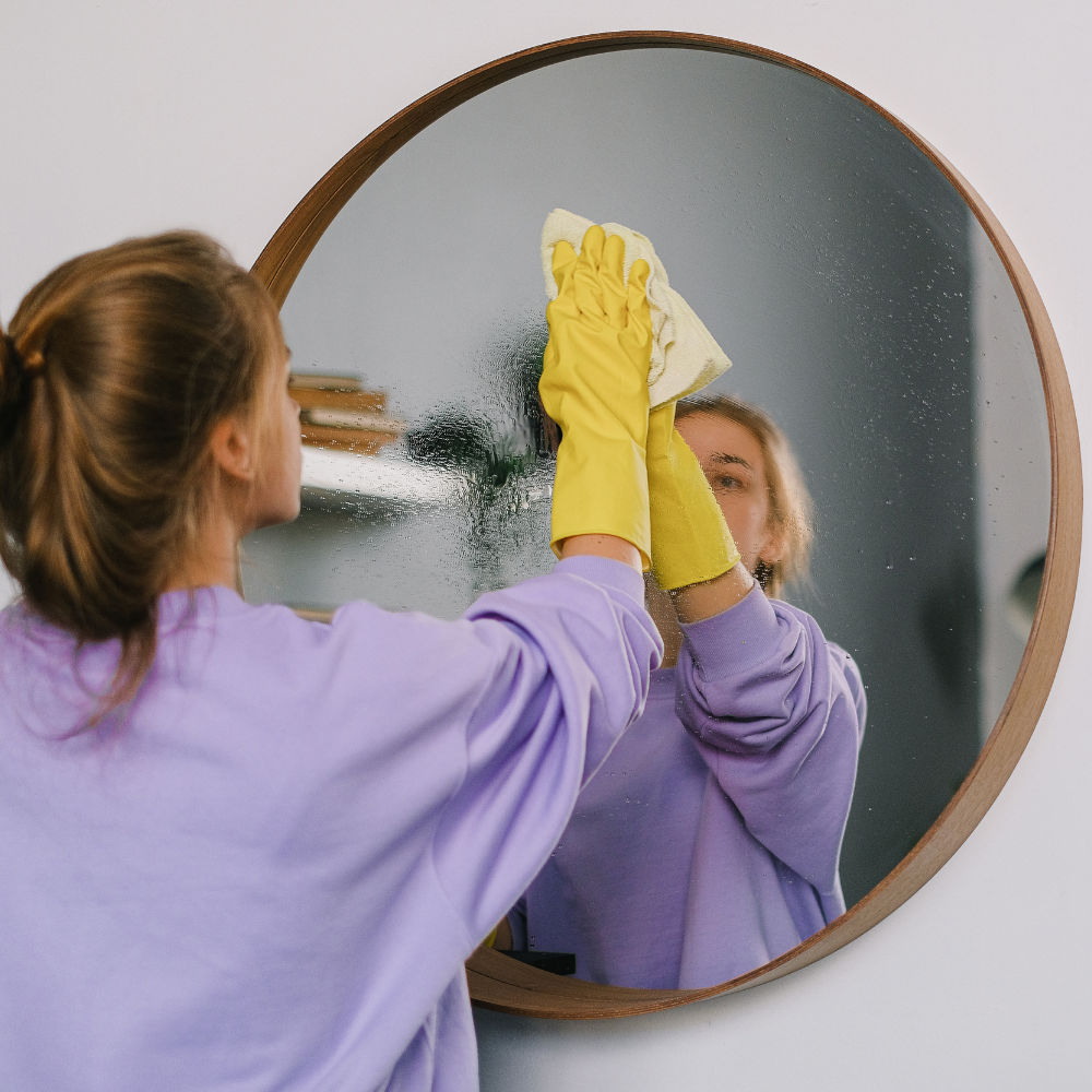 A woman in a lavender sweatshirt and yellow gloves wipes a circular mirror, reflecting her focused expression.