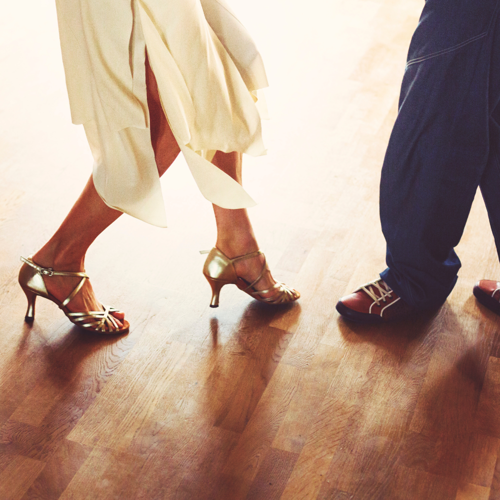 Step Into Confidence C&C #34 Close-up of two dancers’ feet on a wooden dance floor, one in gold ballroom shoes and the other in men’s dance shoes, symbolising confidence and growth.