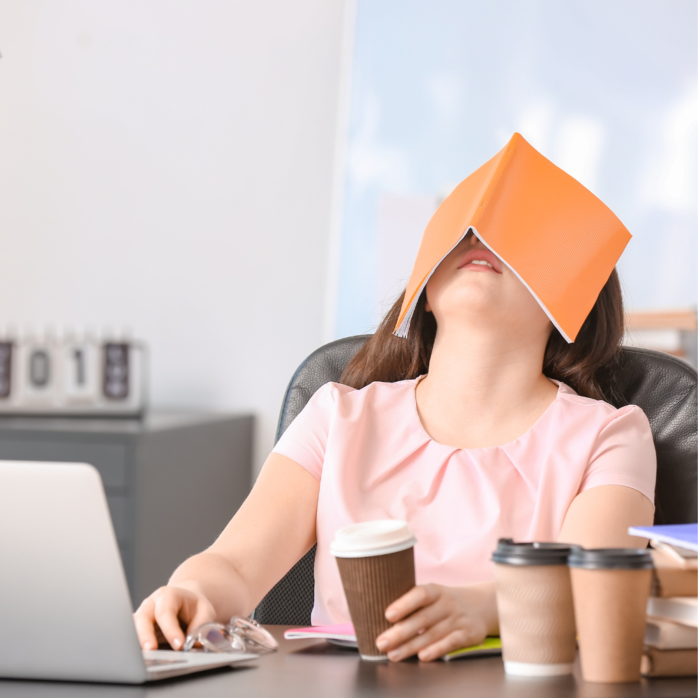A tired woman sits at a desk with a coffee cup and a notebook covering her face, symbolising burnout and exhaustion before starting a wellness reset.