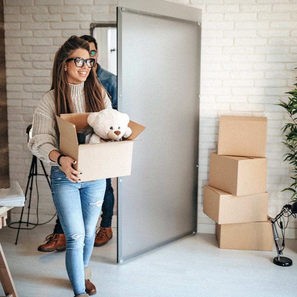 Smiling woman carries a moving box with a teddy bear into a bright new flat; partner follows behind; stacked boxes suggest a fresh start.