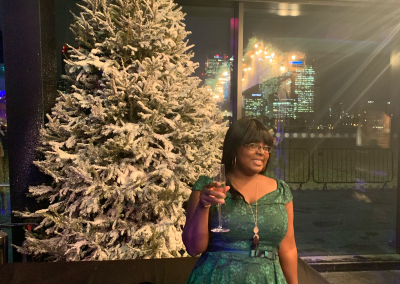 Woman in green dress holding a glass of champagne beside a decorated Christmas tree at night.